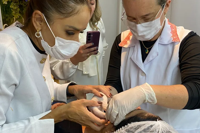 Duas profissionais de máscara aplicando em uma paciente deitada, com as mãos enluvadas sobre o rosto, enquanto uma terceira registra o procedimento