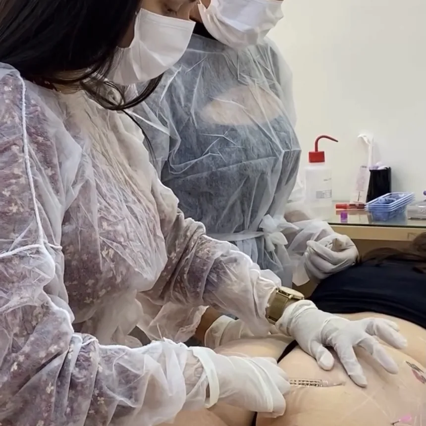 A mentora e uma aluna aplicando em paciente modelo, com mãos enluvadas e a região já marcada